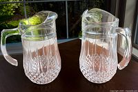 Photo of two clear crystal water pitchers with cut glass base and vertical facets, sitting on a surface near a window with greenery outside.