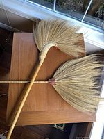 Two decorative brooms from Thailand placed on a wooden table, showing their full length and fan-shaped natural bristles.