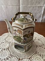 Side view of hexagon shaped tea pot on warming stand showing floral and bird art with gold trim and handle.