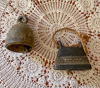 Top-down view of two Thai metal bells on a crocheted decorative cloth: one round with loop top, one rectangular with rope handle and engraved patterns.