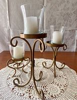 Photo showing all three candle holders with metal scroll stands and glass wind protectors, each fitted with white pillar candles, placed on a lace doily on wooden surface.