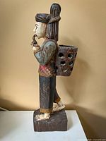 Side view of a hand carved wooden man figure with painted traditional attire, holding a pipe and carrying a basket on his back.