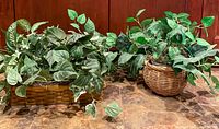 Three silk plants with green leaves each in a different wicker basket planter, arranged on a flat surface against a wood paneled background.