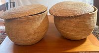 Two large round wicker baskets with unattached lids, showing the shape and size on a wood table.