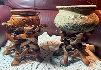 Two bowls each on tripod stands carved from wood. The left bowl is wooden with camels on stand; the right bowl is clay with a plain wooden tripod stand.