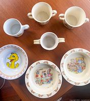 Top-down view of the seven baby mugs and bowls showing design details including Bunnykins and Big Bird themes.
