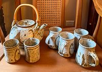 Front view of seven-piece earthenware tea set with teapot, cream pitcher, sugar bowl, and four mugs arranged on a wood chair.