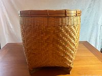 Tall round open top wicker basket showing woven herringbone pattern and slight trapezoidal shape with feet.