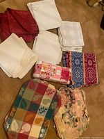 Photo showing folded assorted tablecloths, runners, napkins and hand towels with patchwork, floral and solid colors including red, white, blue, and multi-colored pieces.