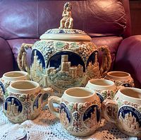 Full set of stoneware tureen with lid and six matching cups arranged on crocheted cloth showcasing castle relief designs