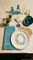 Photo showing two muted green ceramic mugs, teal towel, floral ceramic plate with blue flowers and butterflies, wicker trivets, two blue glass jars with stoppers and rope detail, natural coral, blue beaded coin purse, and a variegated green ceramic dish.