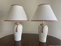 Pair of white ceramic table lamps with cream fabric shades accented with pink trim and tassels, placed on a wood surface against a neutral wall.