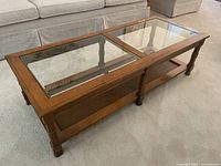 Side angled view of wood and glass coffee table showing clear glass top panels and lower woven wicker shelf, positioned on carpet in front of couch.
