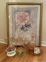 Framed floral abstract artwork in pink tones with four pink stemware glasses, pink glass pitcher on matching plate, multiple votive candle holders and candles arranged in front on floor.