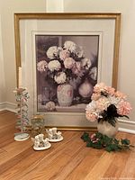 Full view of floral still life framed artwork with gold-tone frame, standing next to candle holders, vase with flowers, faux greenery, and a decorative glass item.