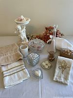 Wide view of collection showing towels, decorative soaps, angel soap dish, artificial flowers, glass bowl with lid, wicker basket, glass jar with bath salts, and other bath accents on table.