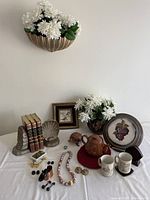 Photo of curated collection on table showcasing white silk flowers, books with ceramic shell bookends, framed bird photo, brown teapot, rustic mugs, jewelry pieces, and figurines.