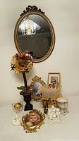 Photo showing oval gold-framed wall mirror, rose topiary, multiple gold frames, swan dish, cherub candle holder, brass mouse figurine, and other decorative items on table.