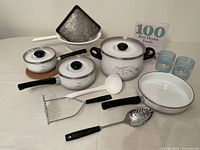 Photo showing the full set of kitchen essentials including three pots/pans with lids, baking pan, food masher, slotted spoon, ladle, two drinking glasses, and cookbook.