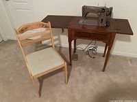 Singer 306K electric sewing machine set up in its dark walnut wooden cabinet sewing table with foldout sides, with light wood chair placed beside it.