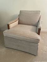 Frontal view of beige fabric traditional armchair with visible removable seat and back cushions, wood paneling on arms, and fabric skirt base.