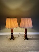 Pair of two traditional style table lamps turned on, showing warm light from pleated woven lampshades and wood and metal bases.