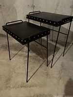 Two black metal TV tray tables with removable black vinyl covered tops, shown standing side by side with visible riveted edges and thin metal legs.