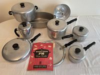 Cookware set arranged on table showing all pots, pans, kettle, lids, and recipe booklet from top view with items separated for clarity
