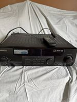 Front of the black Kenwood VR-505 AV receiver showing power cord and remote on top, with multiple buttons and volume knob visible.