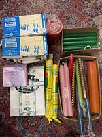 Photo shows various boxed and loose candles arranged on a patterned carpet including Medora Shabbat candles, green taper candles, large red pillar candle and assorted colored taper candles.
