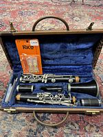 Open view of disassembled clarinet inside blue velvet lined hard case, including barrels, mouthpiece, and bell; unopened Rico clarinet reed pack placed within case