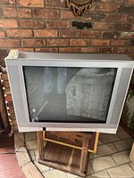 Silver-gray Toshiba CRT TV screen with surrounding bezel and speaker grilles on sides, reflecting some room furniture and brick wall background.