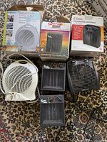 Four electric heaters on patterned carpet including one white fan heater, one boxed Super Furnace ceramic heater, one boxed Classic fan heater, and one black Super Furnace heater.