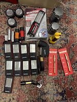 Image showing an assortment of flashlights including multiple Reactor work lights, handheld flashlights with magnets, hanging battery-powered lanterns, a right-angle flashlight, and boxed penlights on a patterned rug.
