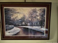 Framed print depicting a winter stream with snow-covered trees and banks in a dark wood frame with white matting.