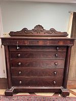 Front view of antique Victorian dresser showing the carved decorative top, five drawers with wooden knobs, and three smaller drawers with keyhole locks, including one key hanging. Dark polished wood with fluted side columns.