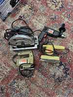 Overview of the power tools: circular saw, drill, two sanders all plugged-in and laid out on a carpeted surface
