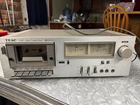 Front view showing cassette compartment, control buttons, VU meters, and control knobs on silver metal faceplate.