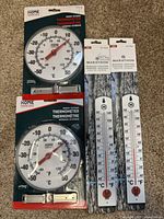 Four sealed thermometers including two Home Essentials round outdoor thermometers and two Marathon vertical strip thermometers on carpeted surface