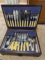 Open wooden box showing vintage cutlery with cream-colored handles including multiple forks, knives, and spoons.