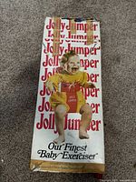 Photo of the original box for the vintage Jolly Jumper baby exerciser with retro design showing a baby in a yellow outfit, the Jolly Jumper logo, and slogan.