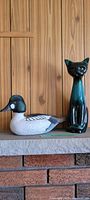 Wooden mallard duck and Blue Mountain Pottery cat figurine displayed on a shelf against wood panel wall.