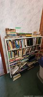 White metal bookshelf fully loaded with books on shelves with loose magazines and papers around bottom
