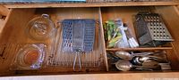Drawer with glass lemon juicer, two clear glass ashtrays, metal flat grater, box grater, small condiment spoons, and yellow and green plastic corn cob holders.