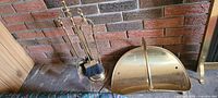 Brass fireplace tool set with stand, showing brush, shovel, poker, and tongs in front of brick wall.