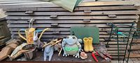 Full view of lot showing metal watering can with floral design, cement frog figurine, rabbit, green plant stands, garden tools and decorative rain boots.
