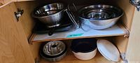 Wide shot of kitchen cupboard showing multiple items: nested mixing bowls, colander, cake pans, ceramic casserole dish