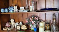 Shelf with multiple decorative items including vintage clocks, blue floral tea set, ceramic figurines, glass candlesticks, floral arrangement, candles, and other small ceramics.