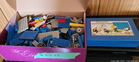 Open pink shoebox filled with assorted Lego pieces including blue plates and wheel parts along with the blue Peanuts pencil case box on the right.