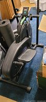 Front and side angle view of Orbitrek elliptical machine on carpeted floor, showing flywheel housing, foot pedals, and digital display.